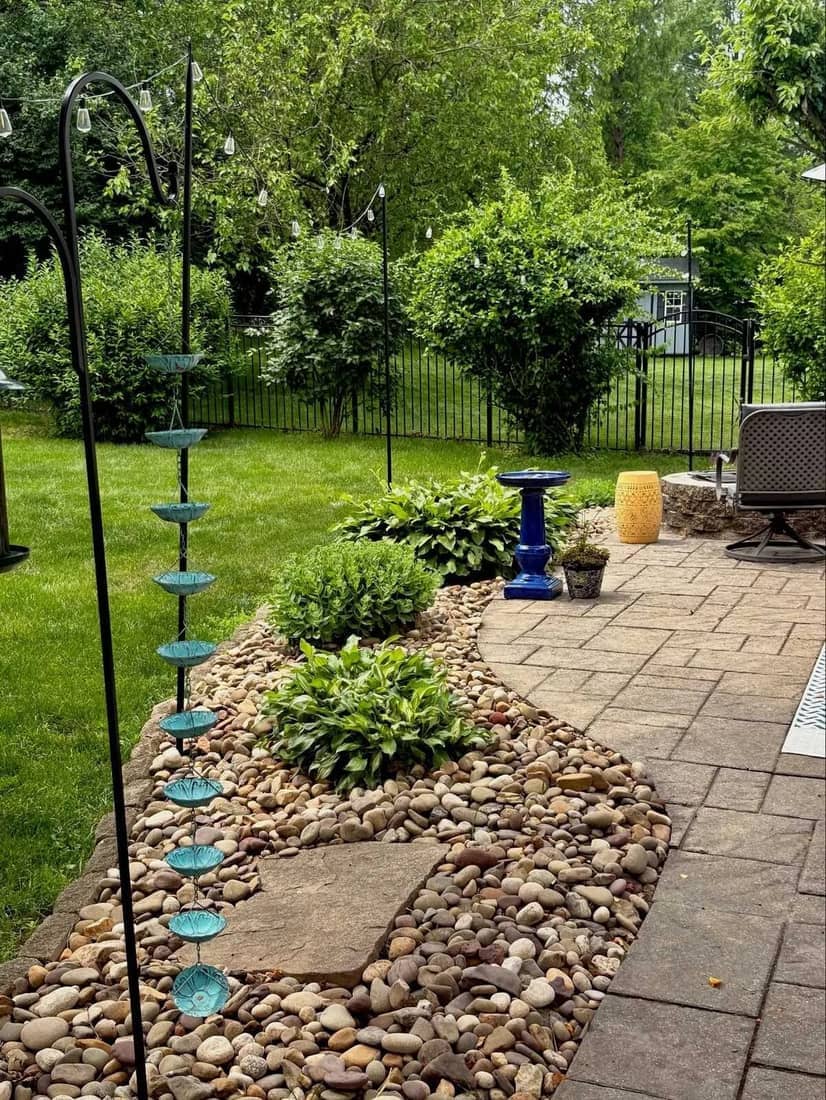 Paver patio edge with river-rock planting bed, hostas, and a rain chain
