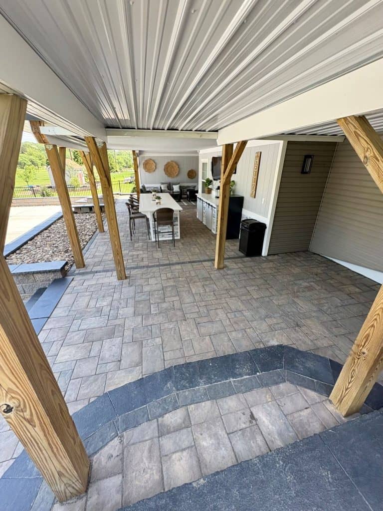 Covered outdoor patio with wood posts, paver flooring, dining area, and built-in kitchen under metal roofing