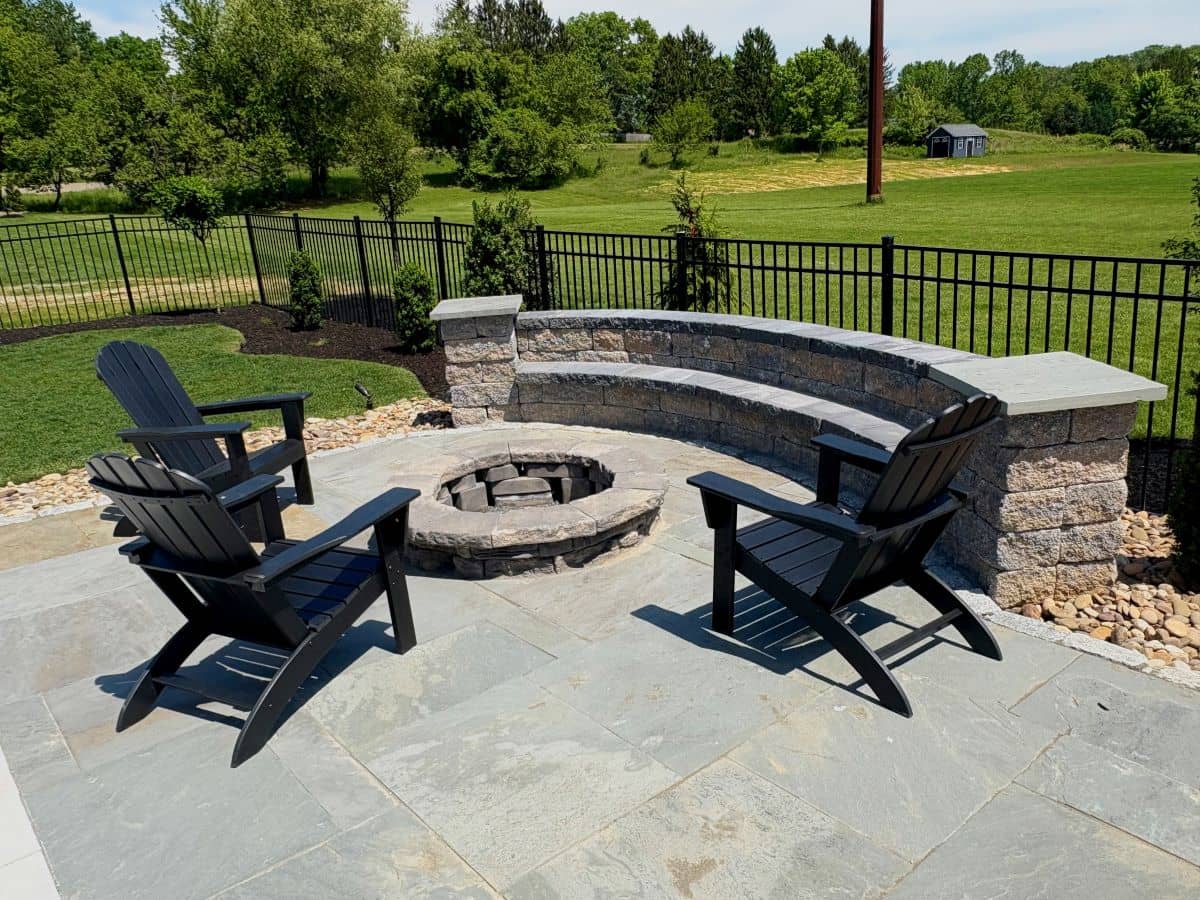Stone patio with a curved bench, round fire pit, and Adirondack chairs facing a fenced lawn